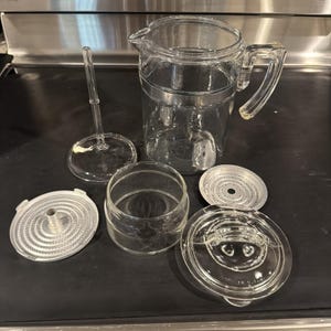 May include: A disassembled clear glass coffee pot with a handle, spout, and various components. The parts include a lid, a filter, a glass cylinder, and a plunger. The coffee pot is designed for brewing coffee.