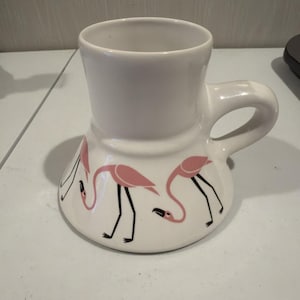 May include: White ceramic mug with a curved base and a handle. The mug features a design of pink flamingos with black legs and beaks. The mug is suitable for hot or cold beverages.