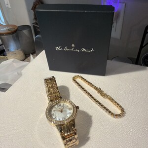 Puede incluir: Un reloj dorado con esfera blanca y bisel con incrustaciones de diamantes, junto a una pulsera dorada a juego con piedras transparentes. Una caja negra con "the Danbury Mint" impresa en el fondo.