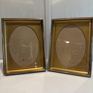 May include: Two rectangular picture frames with a metallic gold finish and a polished sheen. Each frame features a central oval-shaped opening for a photograph, surrounded by a wide, flat border. The frames are standing upright side by side.