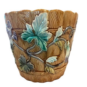 May include: A ceramic planter featuring a brown wood-grain texture with a scalloped rim. The exterior is decorated with raised, glossy relief patterns of green and teal ivy leaves and vines that wrap around the cylindrical body of the pot.
