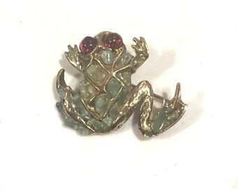 May include: A gold-toned metal frog brooch featuring a textured lattice design filled with small, pale green stone inlays. The frog has two prominent, round, dark red cabochon eyes and a textured surface on its limbs and body.