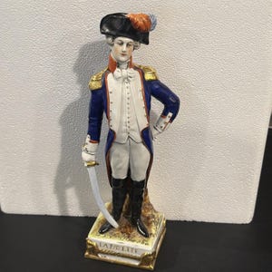 Puede incluir: Figura de porcelana de un hombre con abrigo azul, chaleco blanco y botas negras, sosteniendo una espada. La figura lleva un sombrero negro con una pluma y está etiquetada "LA FAYETTE" en la base. La figura mide aproximadamente 25 cm de altura.