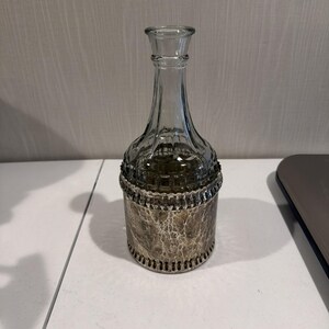 May include: A clear glass bottle with a decorative silver-colored base and rim. The bottle has a long, slender neck and a flared top. The base has a textured, mottled appearance. The bottle is likely for decorative purposes.