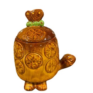 May include: A ceramic turtle-shaped jar with a lid. The jar is brown and yellow with a glossy finish. The lid has a butterfly-shaped handle and green accents. The body of the jar is decorated with floral patterns. A decorative kitchen item.