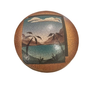 May include: Round decorative object with a landscape scene. The image features a beach with palm trees, mountains, and a blue ocean. The border is a cracked, textured brown.