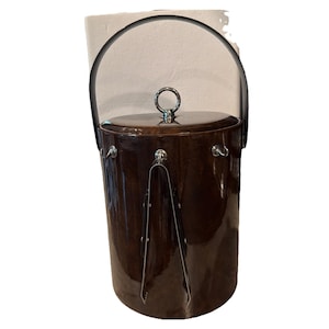 May include: A vintage ice bucket with a dark brown, glossy finish. It features a black handle, a silver-toned lid handle, and silver-toned accents. Stainless steel ice tongs are attached to the front. A classic barware item.