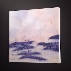 May include: Square art print depicting a landscape scene with figures and kites. The artwork features a soft color palette of purples, pinks, and whites, with figures on hills and kites in the sky. The print is mounted on a white surface.
