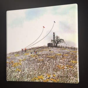 May include: Square ceramic tile featuring a pastoral scene. The image depicts a grassy hill with yellow wildflowers, a windmill, and people flying kites. The sky is a mix of blue and white, and the overall aesthetic is reminiscent of a vintage painting.