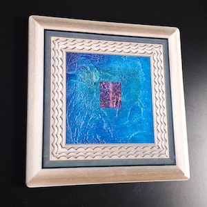 Puede incluir: Obra de arte abstracta enmarcada cuadrada. La obra presenta un fondo azul vibrante con un cuadrado central de tonos morados y rosas. El marco es de color claro con un borde interior ondulado decorativo.