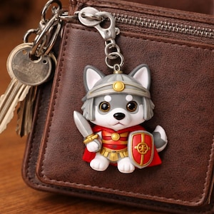 Puede incluir: Un llavero con un lobo guerrero de dibujos animados con casco, capa roja, escudo y espada. El lobo es gris, blanco y marrón claro, con un cierre de llavero plateado. El llavero está sujeto a una cartera de cuero marrón con llaves.