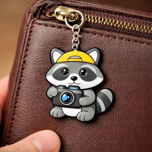 May include: A raccoon keyring wearing a yellow cap and holding a camera. The keyring is grey, white, and black, with a silver chain and clasp. The background is a brown leather wallet.