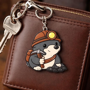 Könnte beinhalten: Ein Schlüsselanhänger mit einem Cartoon-Maulwurf-Charakter. Der Maulwurf trägt einen braunen Bergmannhelm mit gelber Lampe, hält eine Spitzhacke und trägt einen Rucksack. Der Maulwurf ist grau und weiß. Der Schlüsselanhänger ist an einem Schlüssel und einer braunen Ledergeldbörse befestigt.