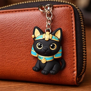 May include: A black cat keychain with a gold and turquoise Egyptian-themed design. The cat has large, expressive eyes and is attached to a silver chain. The keychain is hanging in front of a brown leather wallet.