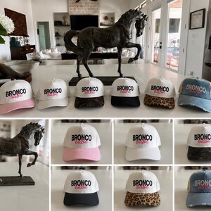 May include: A collection of baseball caps featuring the text BRONCO babe in black and pink lettering. The caps come in various styles, including solid white, pink, black, leopard print, snakeskin print, and distressed blue denim with mesh backs.