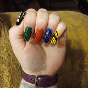 Puede incluir: Una mano que muestra uñas artificiales largas, coloridas y brillantes. Las uñas están pintadas en verde, rojo, azul y amarillo, con rayas diagonales plateadas. La uña del pulgar es negra con un diseño blanco. Se ve una correa de reloj morada en la muñeca.