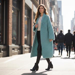 May include: A light blue, long, open-front crochet coat. The coat is knee-length and has a shawl collar. The person is wearing black pants and black ankle boots. The background is a city street with brick buildings and pedestrians.