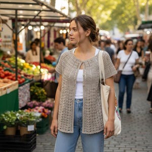 May include: A beige, short-sleeved crochet cardigan with a button closure. The cardigan is worn over a white top and paired with blue jeans. A canvas tote bag is carried over the shoulder. The background shows a bustling outdoor market.
