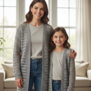 May include: Two people wearing matching gray open-weave cardigans, gray t-shirts, and blue jeans. The cardigans have a long, flowing design. The background includes a window, a couch, and a potted plant.