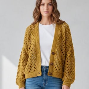以下が含まれることがあります： マスタードイエローのオープン織りのカーディガン。3つの茶色のボタンが付いています。カーディガンは長袖で、白いTシャツとブルージーンズの上に着用されています。スタイリッシュで快適な衣服です。