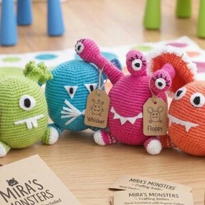 May include: Four colorful, handmade crochet monster toys. The monsters are green, blue, pink, and orange, each with unique features and tags. The tags have names like "Gordo" and "Whisker". The toys are displayed with a box that says "Mira's Monsters".