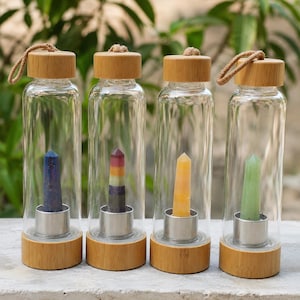 May include: Four clear glass water bottles with bamboo lids and carrying loops. Each bottle contains a single vertical crystal point secured in a silver-toned metal base. The crystals are blue, multicoloured, yellow, and light green in colour.