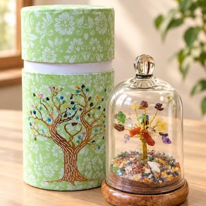 Könnte beinhalten: Eine zylindrische hellgrüne Geschenkbox mit weißem Blumenmuster und Baumillustration neben einer Glaskuppel, die eine Drahtbaumskulptur mit bunten Edelsteinblättern auf einem runden Holzsockel mit einer Schicht aus kleinen Steinen enthält.