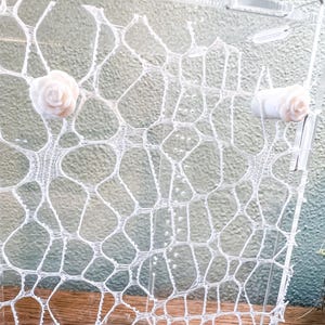 May include: A clear acrylic display stand features a white, web-like, open-work lace pattern across its surface. Two small, pale pink, three-dimensional rose-shaped embellishments are attached to the upper left and right sides of the panel.