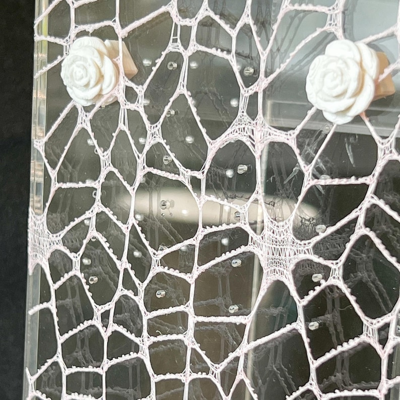 May include: A clear acrylic display stand featuring a pale pink, web-like lace overlay. Two white, three-dimensional rose-shaped embellishments are attached to the upper portion of the lace, which is secured against the transparent surface.