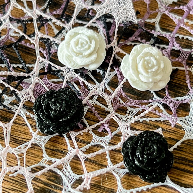 May include: Two white rose-shaped buttons and two black rose-shaped buttons arranged on a white and black web-like lace fabric. The buttons feature detailed, layered petals and a smooth, glossy finish, contrasting with the intricate, open-work texture of the underlying lace material.