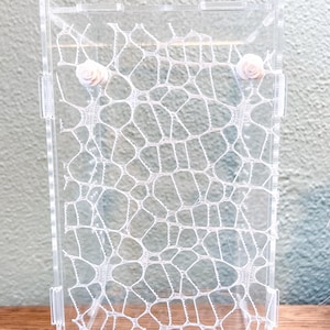 May include: A clear acrylic display case featuring a white lace panel with an intricate, web-like pattern. Two small, pale pink rose-shaped embellishments are attached to the upper portion of the lace, positioned symmetrically on the left and right.