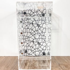 May include: A clear acrylic rectangular display case featuring a black web-like lace pattern on the interior back panel. The structure includes a vertical side panel with four small circular openings and a single light-colored circular knob at the top.