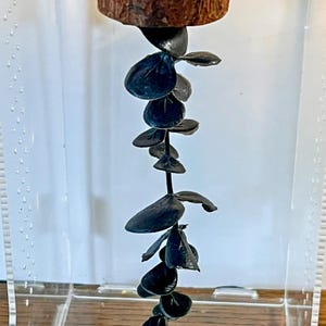 May include: A vertical decorative piece featuring a dark, circular wooden base at the top, from which a long, slender stem hangs downward. The stem is adorned with numerous small, rounded, dark-colored leaves arranged in a cascading pattern.