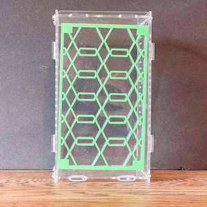May include: A rectangular clear acrylic enclosure featuring a light green geometric lattice pattern on the front panel. The structure is assembled with visible interlocking tabs and slots along the sides, creating a transparent box design.
