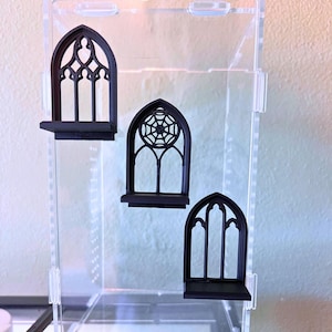 May include: Three black, 3D-printed gothic window-shaped shelves attached to a clear acrylic display case. Each shelf features a pointed arch frame, with one center piece displaying a spiderweb design and two side pieces showing arched mullions.