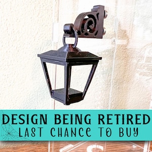 May include: A black miniature wall-mounted lantern with a scrollwork bracket, featuring a tapered frame and a square base. The image includes the text: DESIGN BEING RETIRED LAST CHANCE TO BUY. The lantern is attached to a clear acrylic panel.