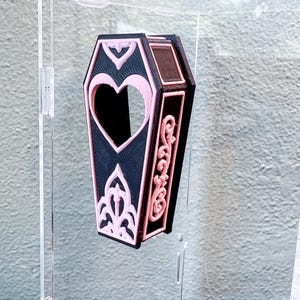 May include: A black coffin-shaped container featuring light pink decorative trim. The lid displays a heart-shaped cutout at the top and an ornate, symmetrical filigree pattern at the bottom. The side panel is adorned with a raised, scrolling vine design.