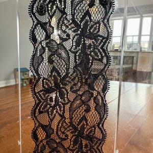 Op de afbeelding: Een decoratief paneel met ingewikkeld zwart kant met bloemmotieven. Het kant is geplaatst in een helder acryl frame, met kleine zwarte roosvormige versieringen aan de bovenkant. Het ontwerp is zichtbaar tegen een vage achtergrond.