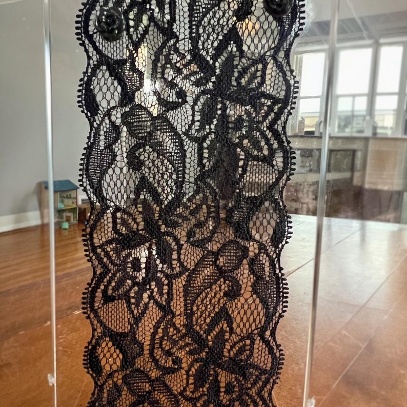 Op de afbeelding: Een decoratief paneel met ingewikkeld zwart kant met bloemmotieven. Het kant is geplaatst in een helder acryl frame, met kleine zwarte roosvormige versieringen aan de bovenkant. Het ontwerp is zichtbaar tegen een vage achtergrond.