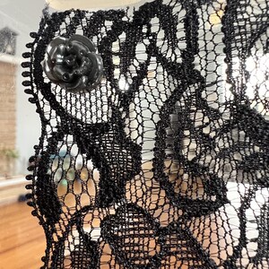 Op de afbeelding: Close-up van zwarte kanten stof met een bloemenpatroon. Een zwarte, roosvormige knoop is aan het kant bevestigd. Het kant heeft een delicate, open weefsel, waardoor een doorschijnend effect ontstaat. De algehele esthetiek is elegant en ingewikkeld.