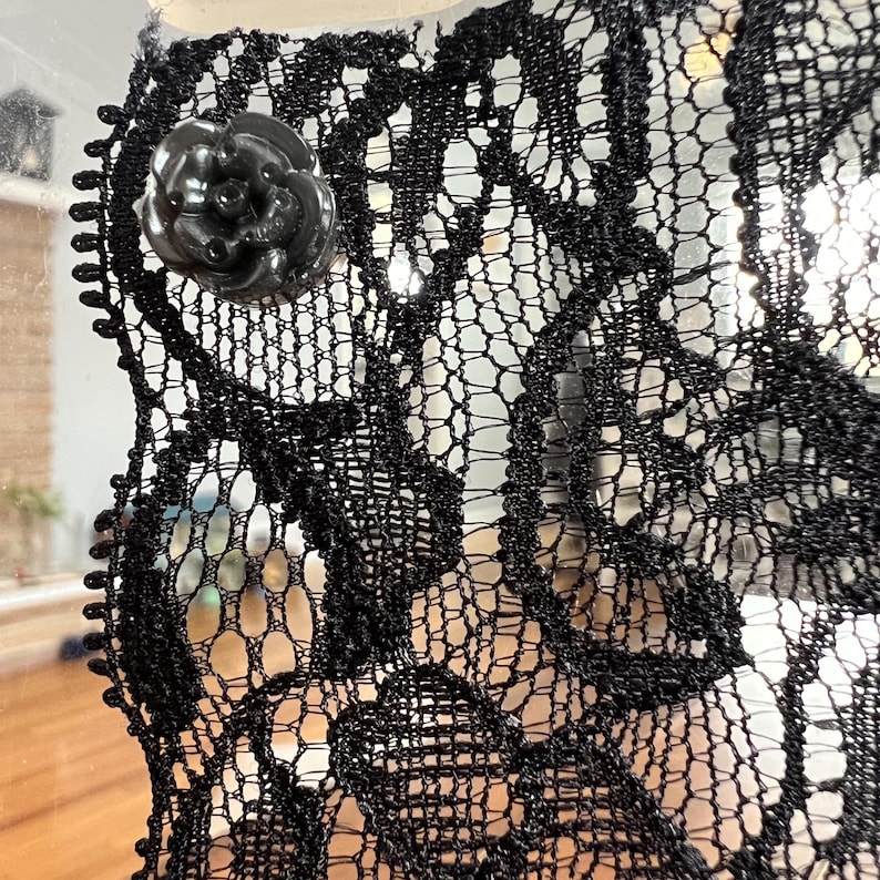 Op de afbeelding: Close-up van zwarte kanten stof met een bloemenpatroon. Een zwarte, roosvormige knoop is aan het kant bevestigd. Het kant heeft een delicate, open weefsel, waardoor een doorschijnend effect ontstaat. De algehele esthetiek is elegant en ingewikkeld.