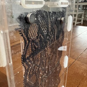 Op de afbeelding: Een heldere acryl display case met een zwarte kanten inzet. Het kant wordt op zijn plaats gehouden door kleine zilveren magneten. De behuizing heeft een rechthoekige vorm en is ontworpen om delicate items te presenteren.