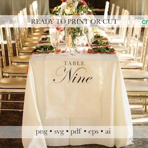 May include: A wedding reception table setting with a cream-colored tablecloth and gold chairs. The table is set with plates of food, glasses, and floral centerpieces. The tablecloth has the words "Table Nine" printed on it.