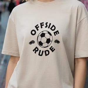 Op de afbeelding: Beige T-shirt met de tekst "OFFSIDE RUDE" rondom een voetbalafbeelding. Twee kleine schoenafbeeldingen flankeren de bal. Het shirt is een casual stijl met korte mouwen.
