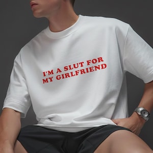 May include: White crew neck t-shirt with the red text "I'M A SLUT FOR MY GIRLFRIEND". The shirt is short-sleeved and has a relaxed fit. The person is wearing black shorts and a silver watch.
