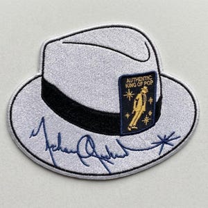 May include: A white fedora hat patch with a black band and a rectangular navy blue emblem featuring a gold silhouette of a dancer and the text AUTHENTIC KING OF POP. A dark blue embroidered signature is visible across the lower brim area.