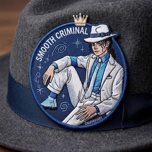 May include: A dark gray felt fedora hat featuring a circular embroidered patch. The patch depicts a figure in a white suit and fedora with a blue shirt and tie, labeled SMOOTH CRIMINAL with a small crown icon above and CHAPPEYVARY below.