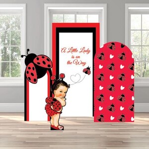 May include: Ladybug-themed party decorations. The scene includes a ladybug cutout, a door frame with the text "A Little Lady is on the Way", and a ladybug-patterned arch. The color scheme is red, black, and white.