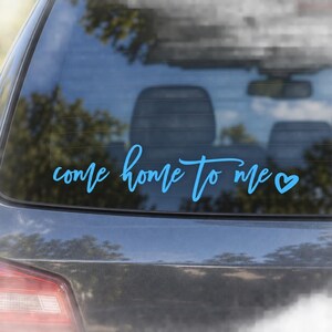 May include: A light blue vinyl decal applied to a car rear window. The decal features the text "come home to me" written in a flowing, cursive script font, followed by a small, simple heart symbol at the end of the phrase.