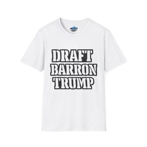 May include: White t-shirt with the text "DRAFT BARRON TRUMP" in bold, black capital letters. The shirt has a crew neck and short sleeves. The brand logo is visible on the upper chest.